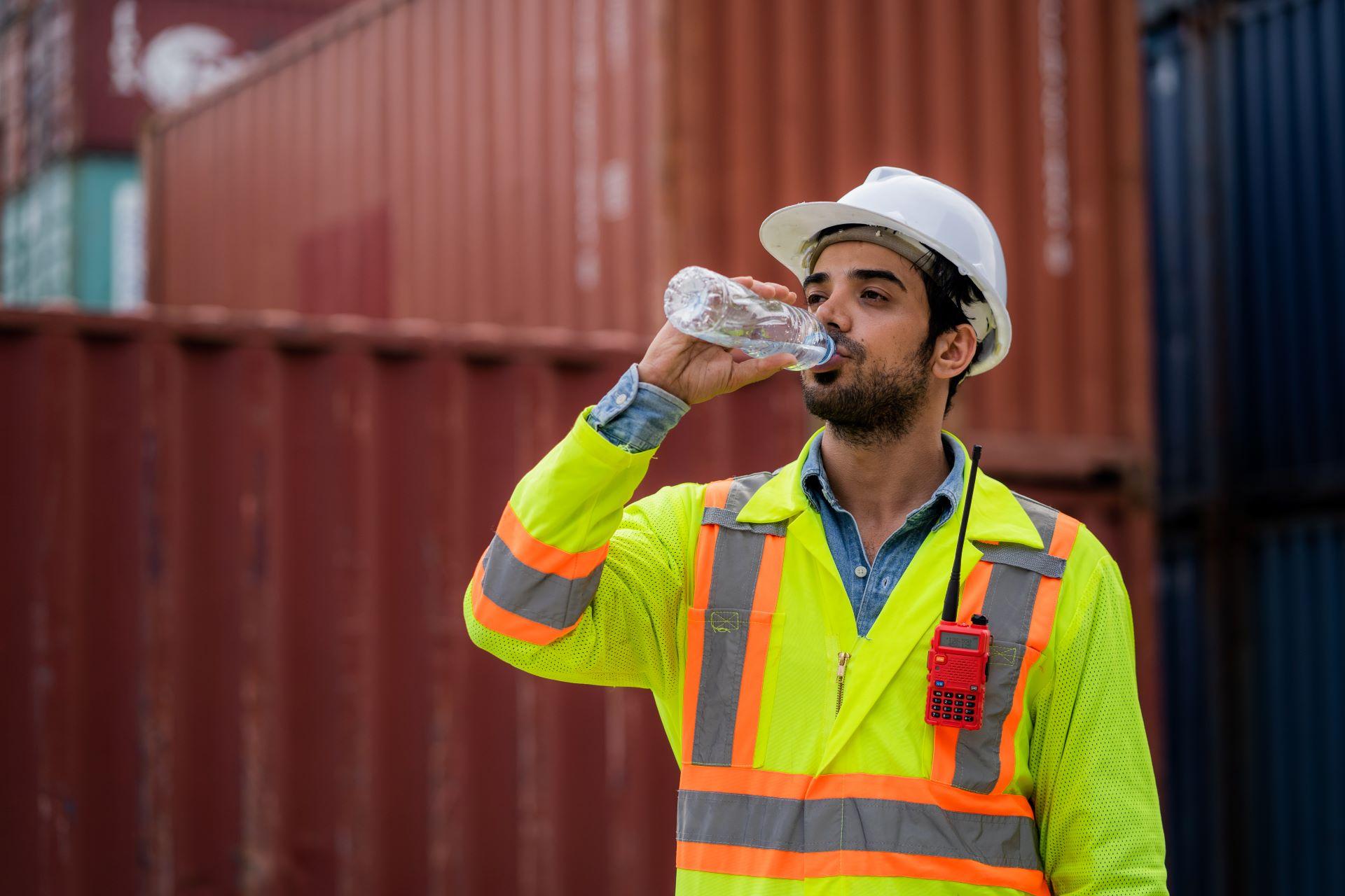 in Bauarbeiter in einer Sicherheitsweste und einem Schutzhelm trinkt Wasser aus einer Plastikflasche. Im Hintergrund sind Container zu sehen, die eine industrielle Umgebung andeuten.